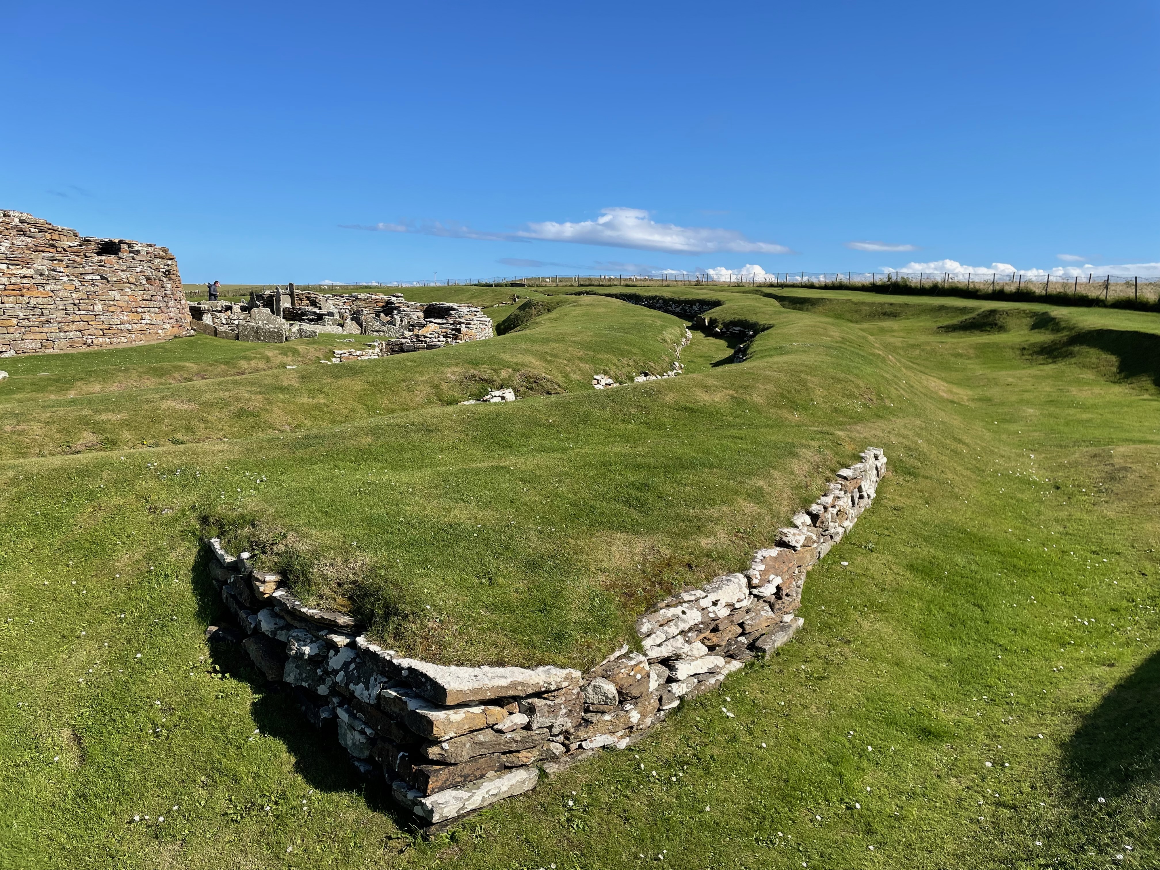 Side of a round building made from flat stones. Concentric banks and ditches surround it, shored up with stone walls. Remains of low buildings in stone visible next to it in the innermost circle.