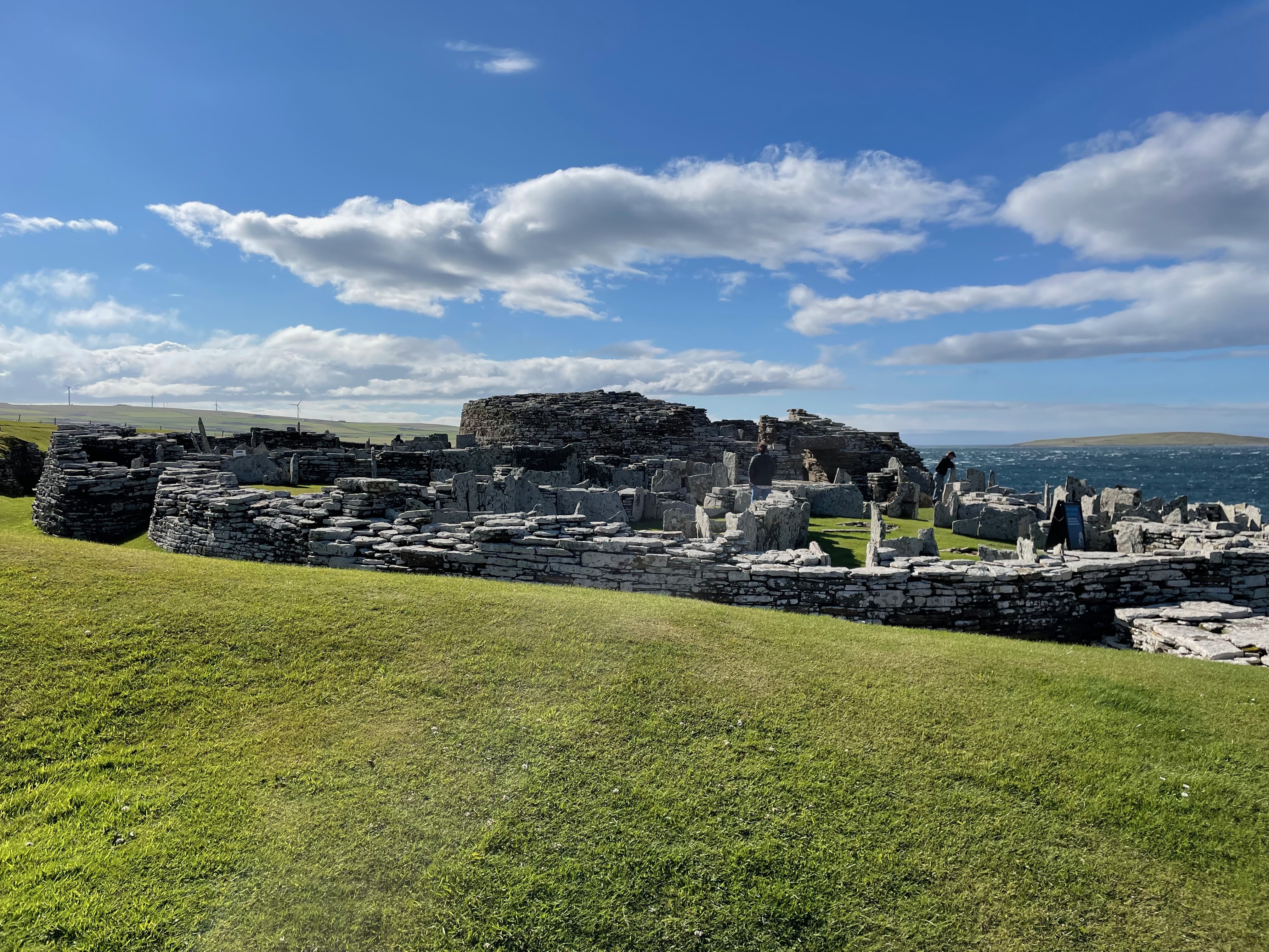 Low stone wall forming what loos to be a circle, thin upright stone slabs are visible within and the remains of a taller round building are across.  Bright blue sky with clouds.  The sea to the right.  View from a raised bank.