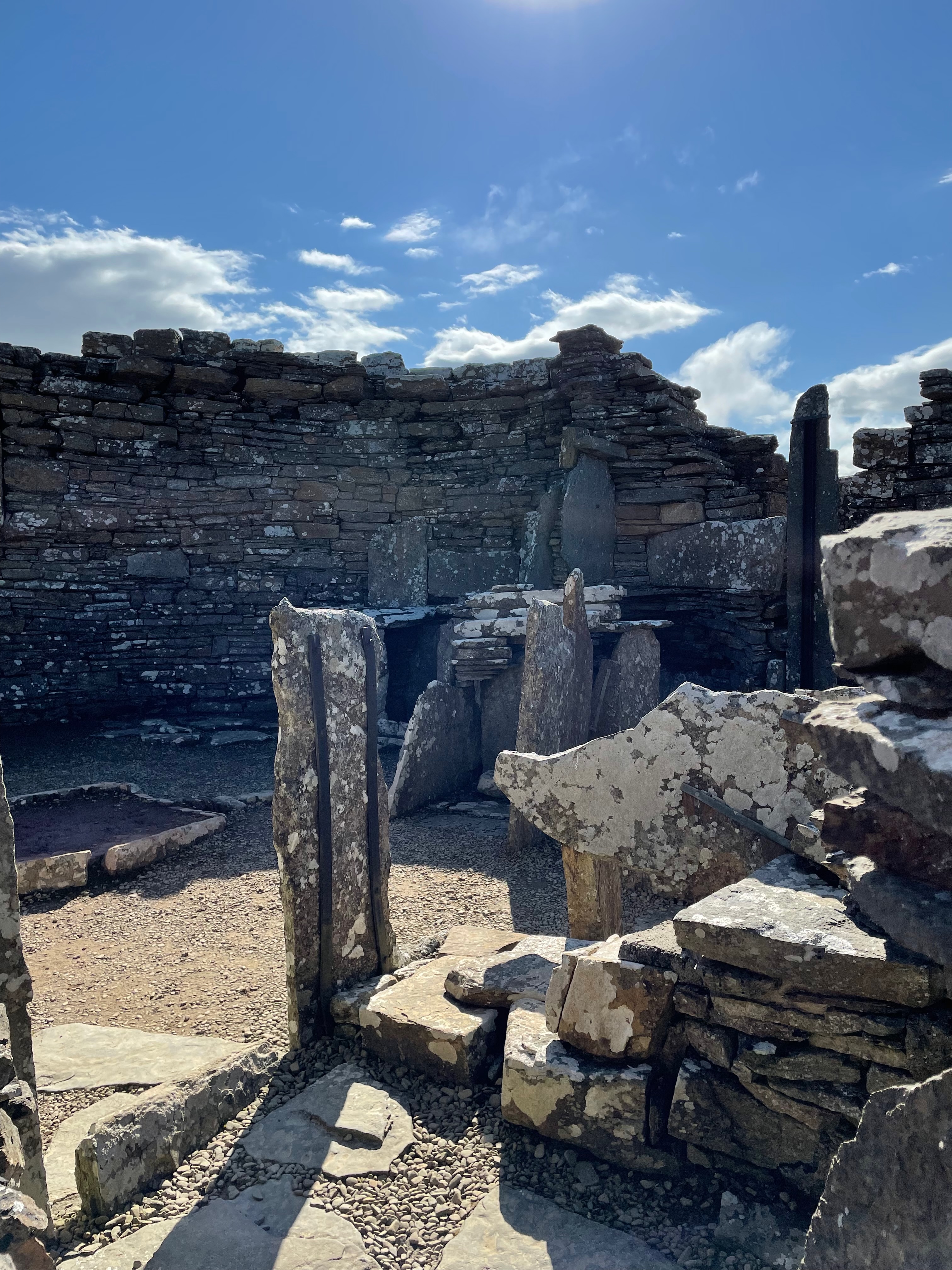 A hish stone wall, slightly curbed. Before it is a collection of thin upright stones, another wall and slow thin stones set in the earth marking what look like room boundaries and a hearth.