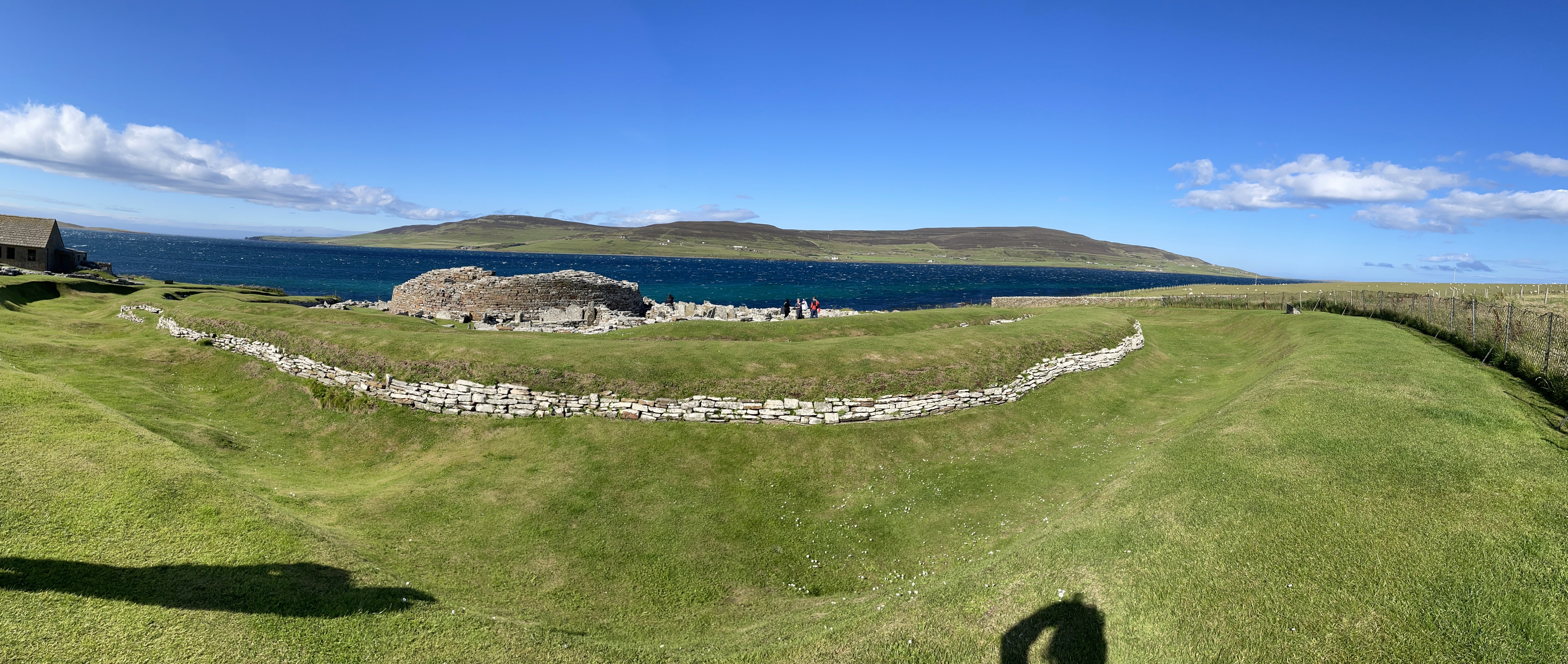 Panoramic shot of a ruined round stone building, with low stone walls arround it, a grassy bank around that with stone walls showing, a ditch around that and then another grassy bank.  The shadow of the photographer is visible on this bank.  Beyond is the sea and another island.