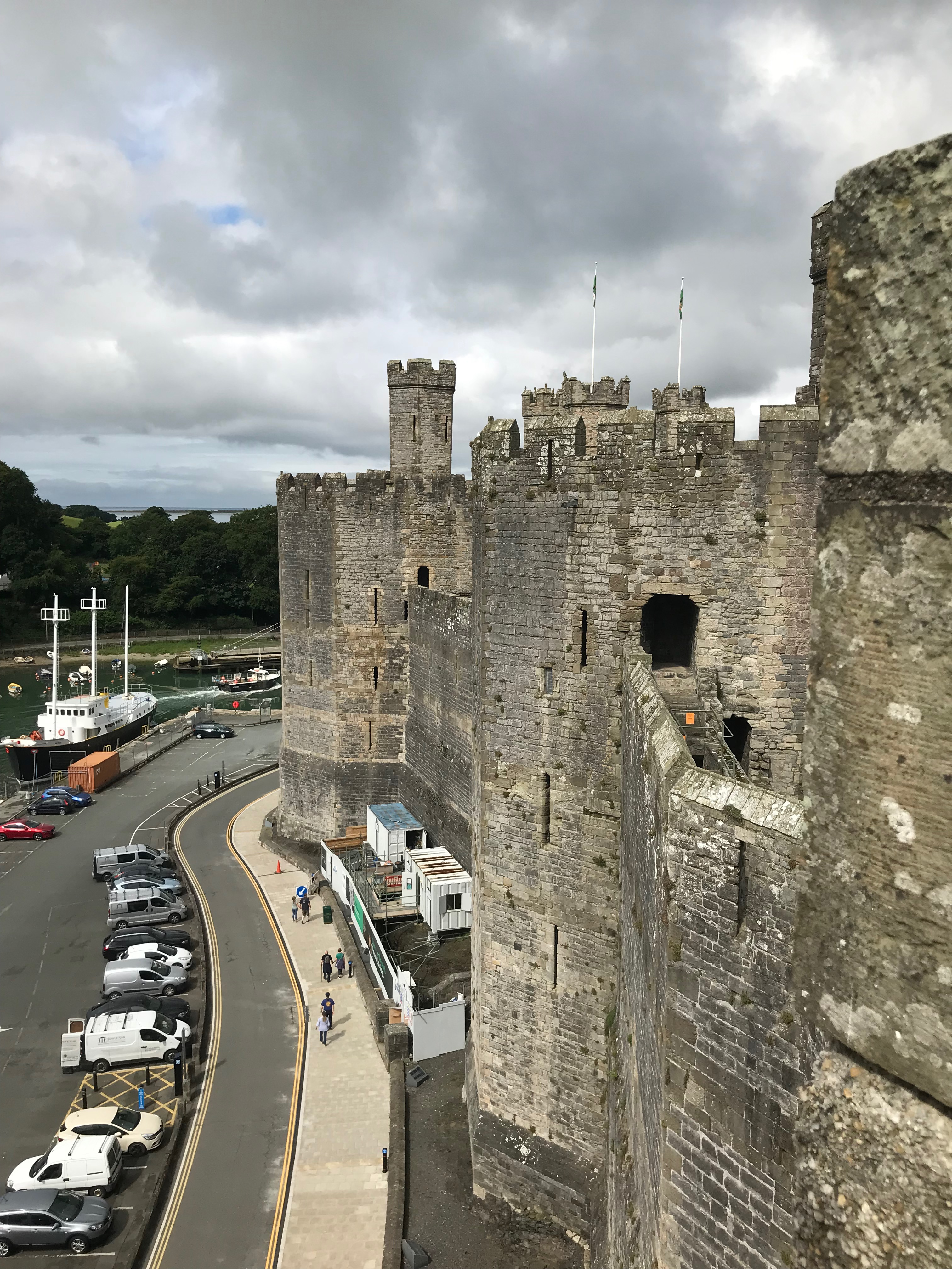 The walls of a castle with hexagonal turrets, buy a car park in front of a river.