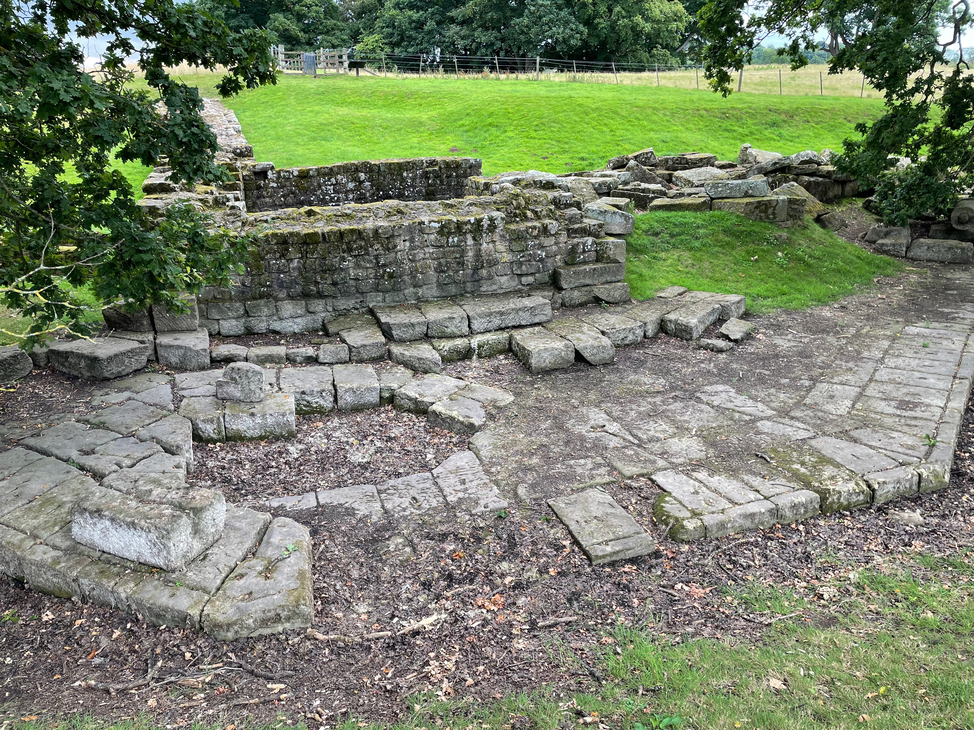 The remains of a possibly hexagonal base in stone with a stone wall behind it and a small chamber visible.