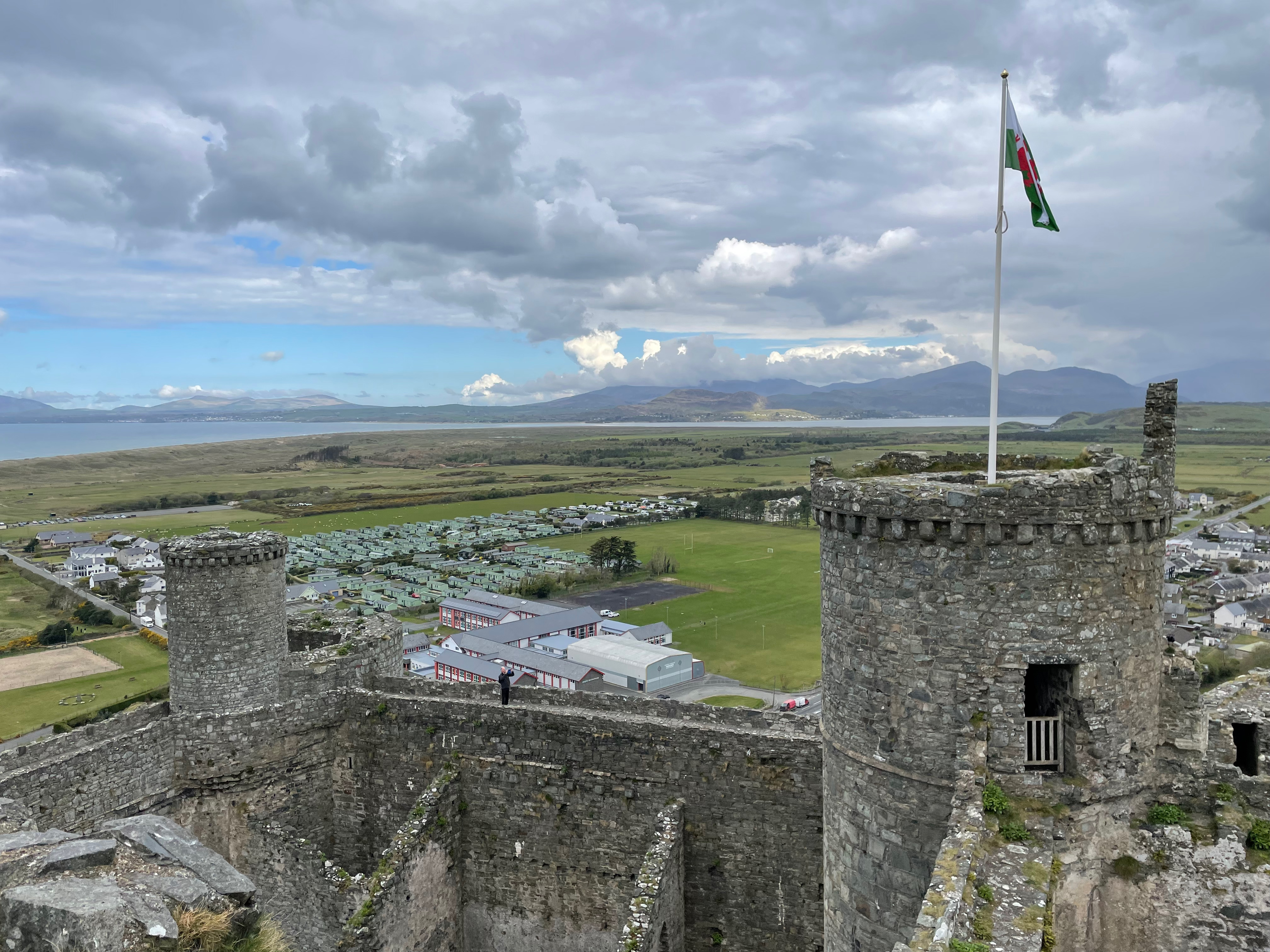 View from on high of a round castle turret flying a flat and then a wall joining it to a second current.  Estuary and mountains in the distance.  Presumably taken from another turret.