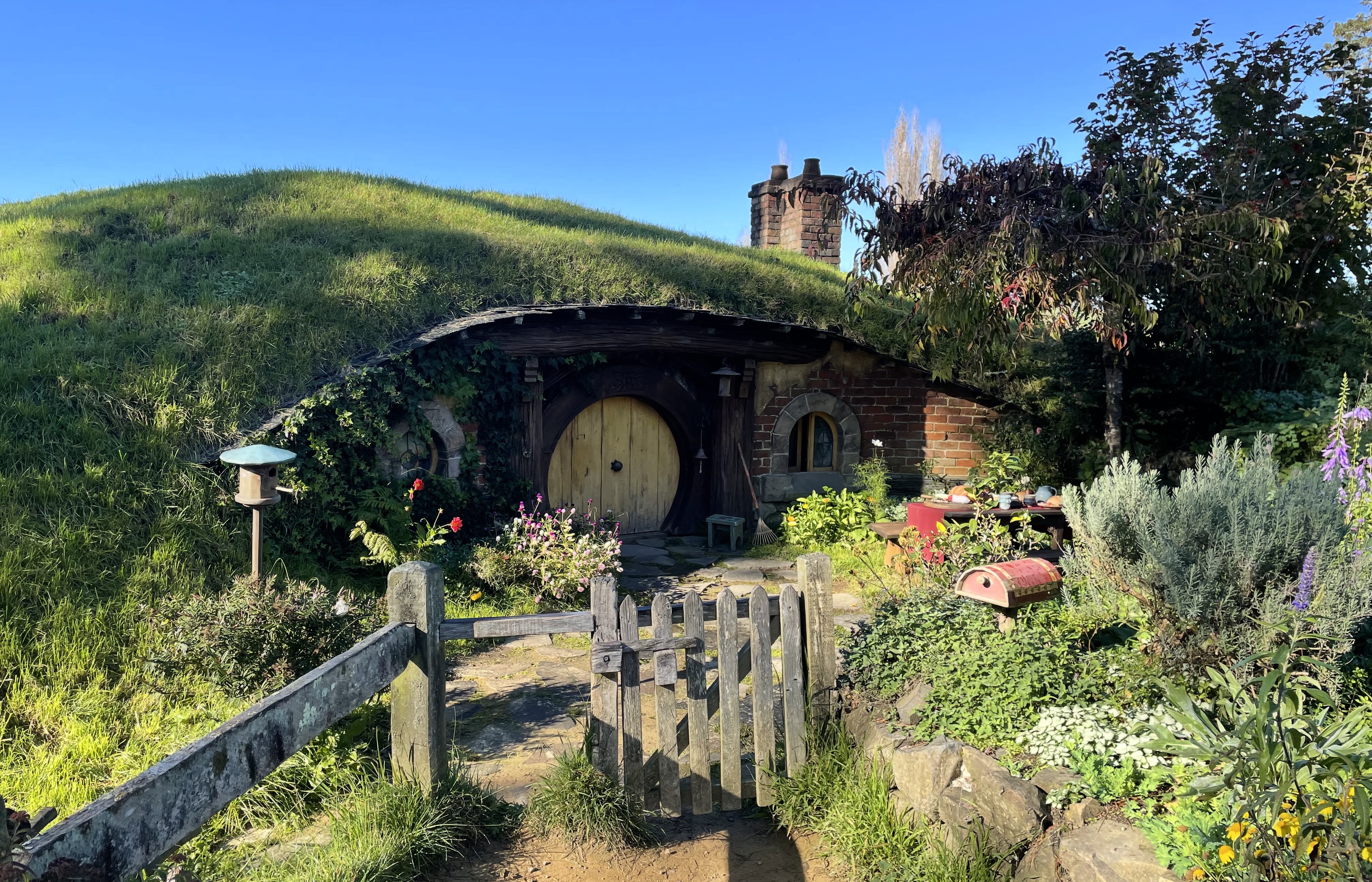 A hobbit hole set into a low green mound. A chimney is visible. It has a yellow door and two windwos. There is a bird nesting box with a green roof and a perch to one side. A table set with food is in the front garden. There is a mailbox beyond the gate.