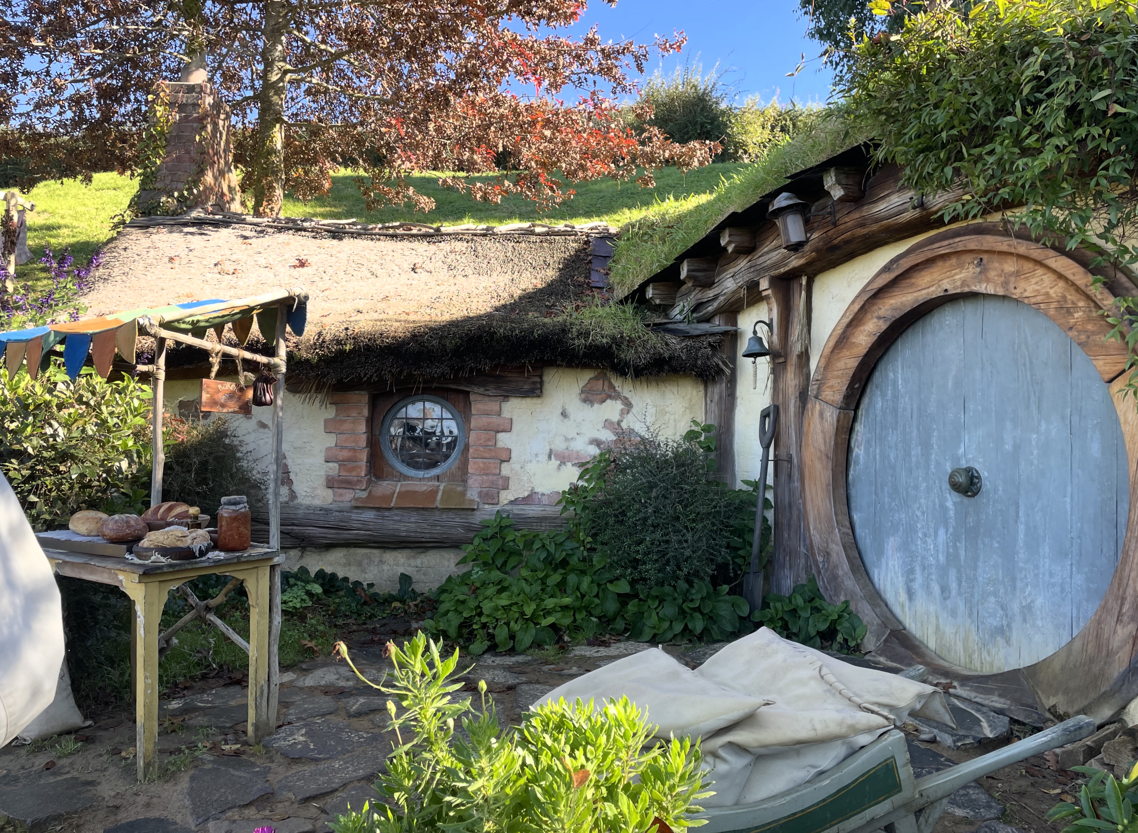 A hobbit hole with a round blue door on the right.  Facing is a small round window in the wall.  Before the door a wheelbarrow loaded with sacks can just be seen behind a bush.  To the left of the window is a small stall with coloured bunting displaying loaves of bread and a jar of some kind of preserve.