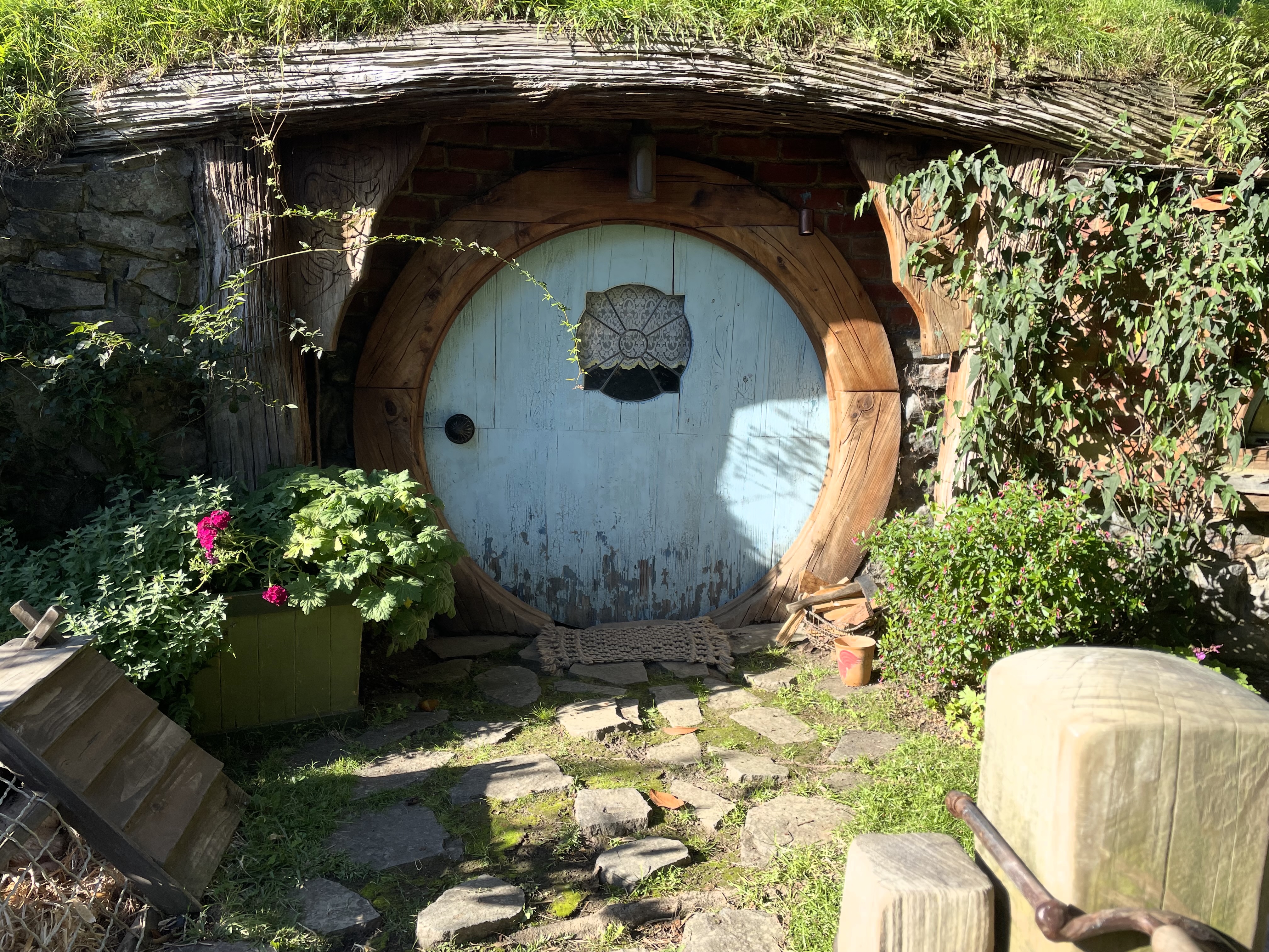 A hobbit hole with a blue door withe a circular paned window in it and a lace curtain bisible behind.