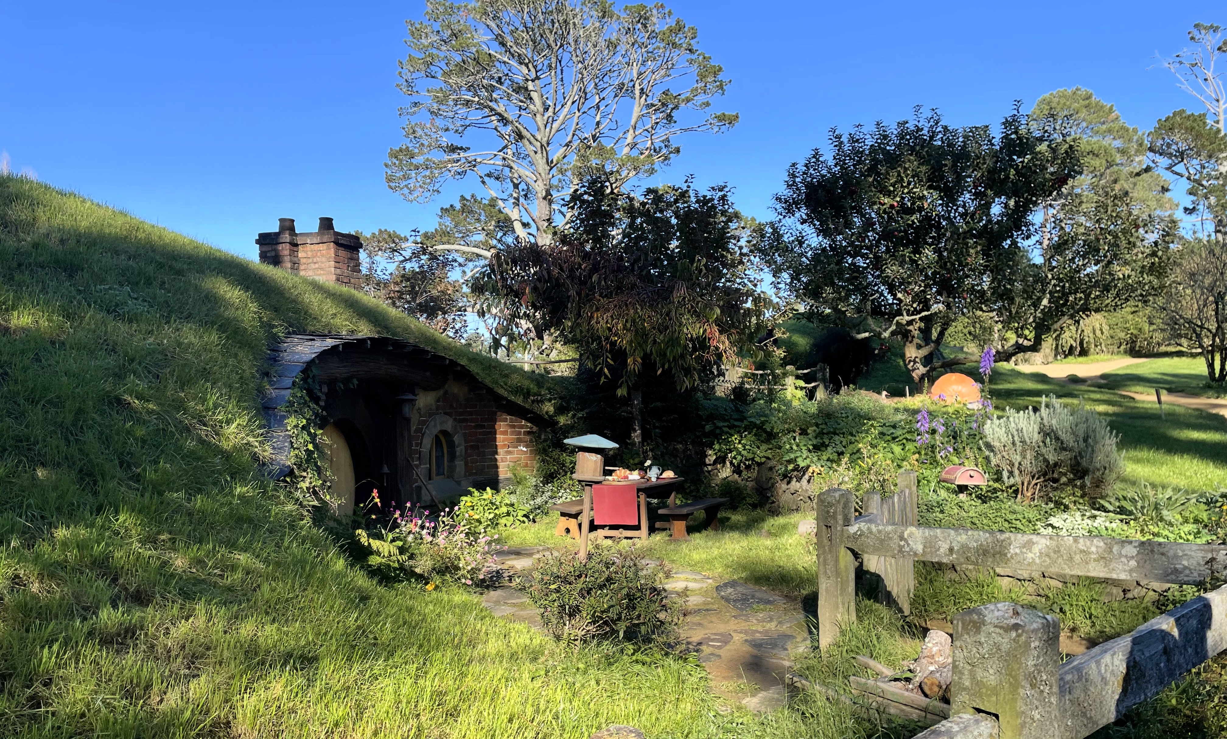 A sloping hill with chimneys and the shady entrance to a hobbit hole.  We are looking across to it from a fence.  In the front garden is a tresle table with benches, spread with food.  A small bird circulat bird box on a stand is next to it.  In the field beyond is a giant orange pumpkin.