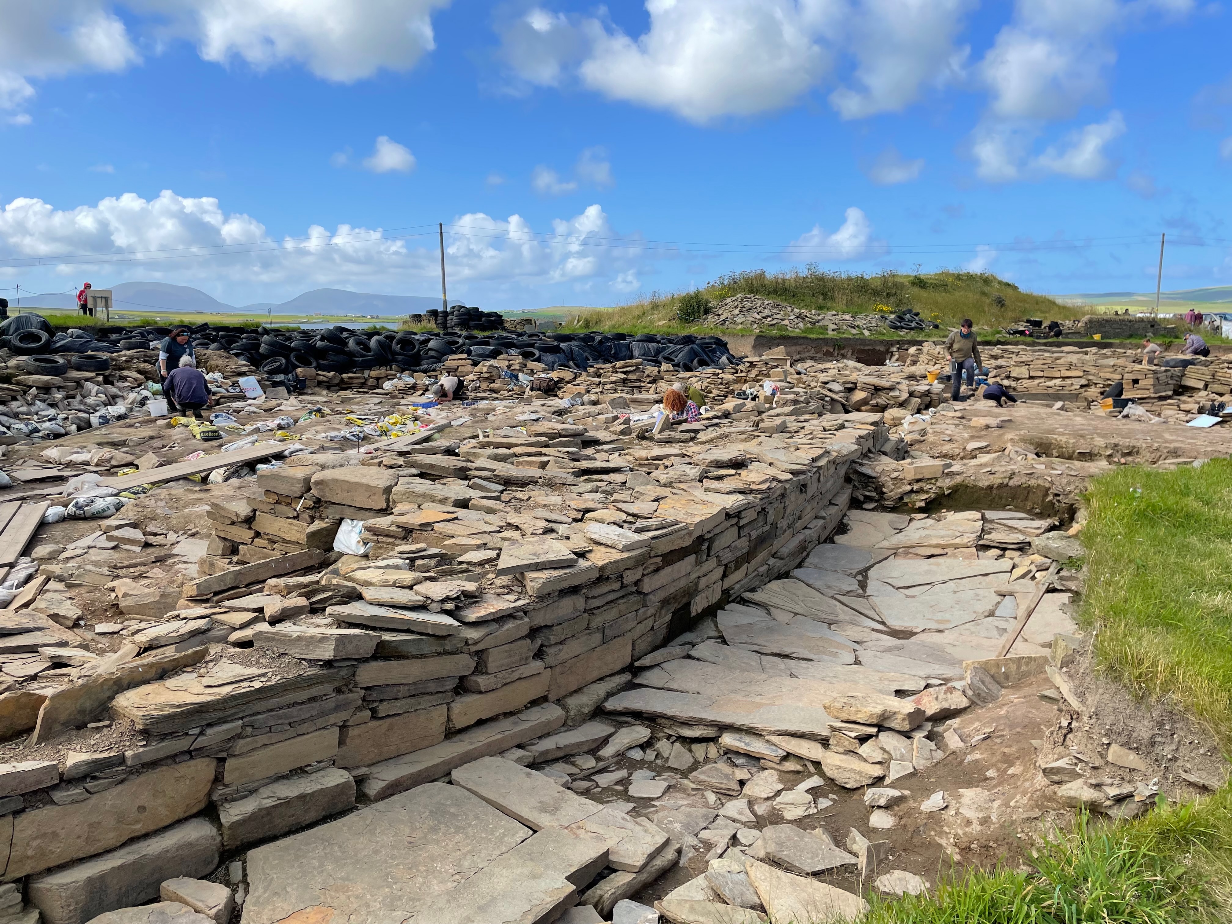 A curved wall, a couple of feet high, made of flat stones and a flagged path in front of it.  Archeologists are digging.  In the background tyres are piled up and there is grass beyond.