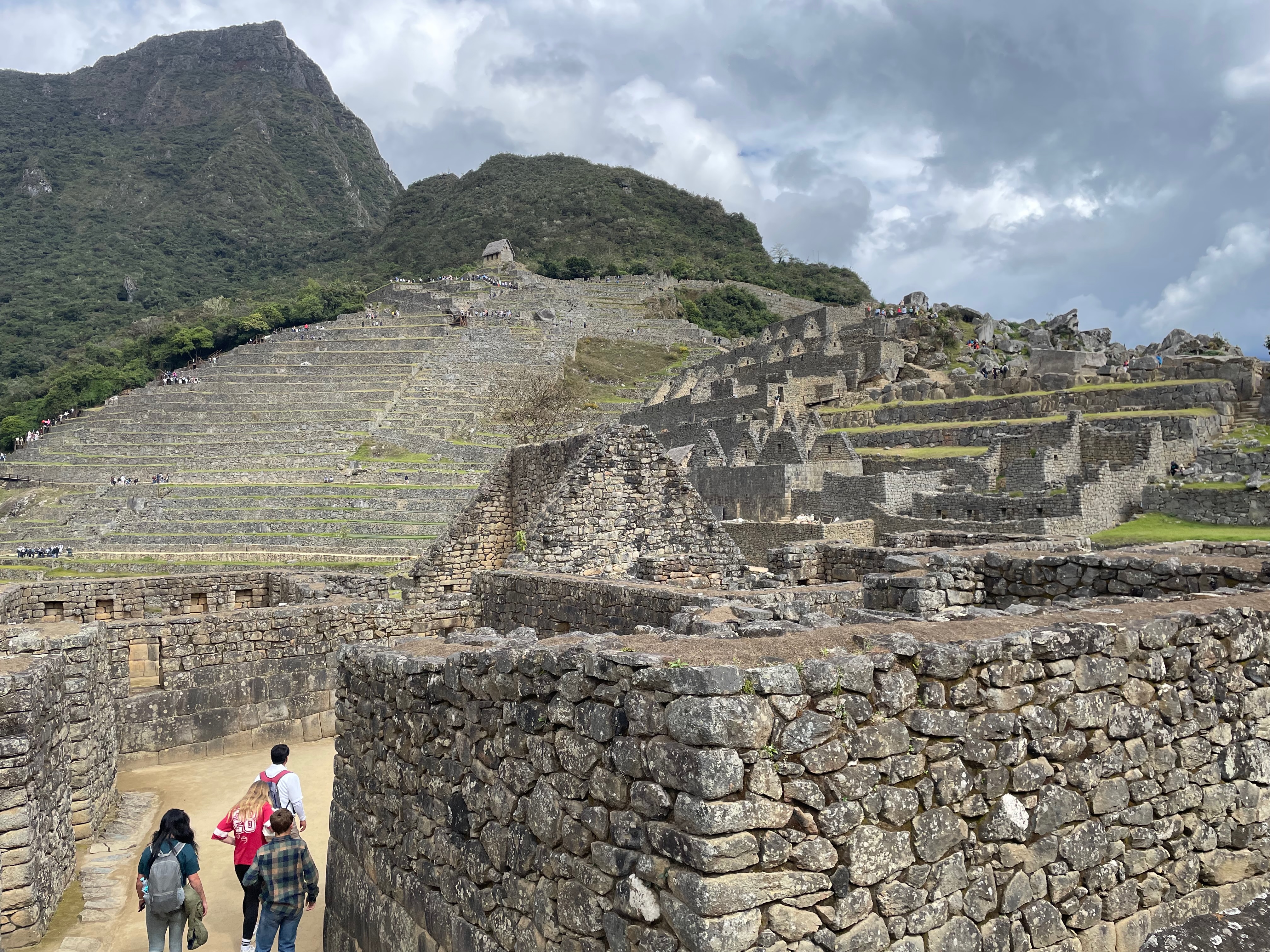 High stone wall leading to a courtyard space.   On the right the remains of walled building proceed up a slope.  Directly ahead is an entirely terraced slopw.  A Mountain is beyond.