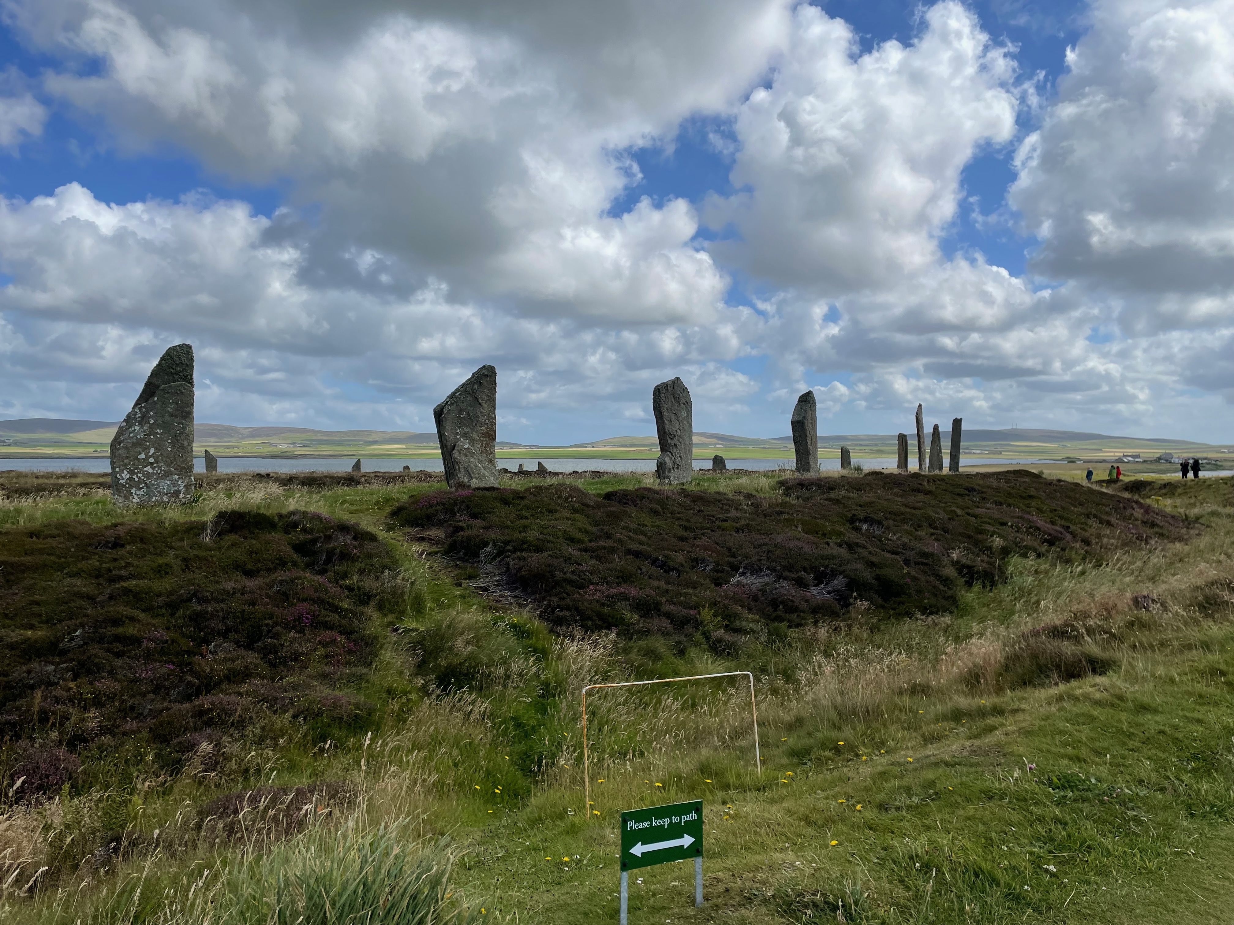 Half a stone circle - a line of stones nearby across a ditch and then curving round so smaller stones can be seen behind.  Against a blue cloudy sky, hills and a body of water.