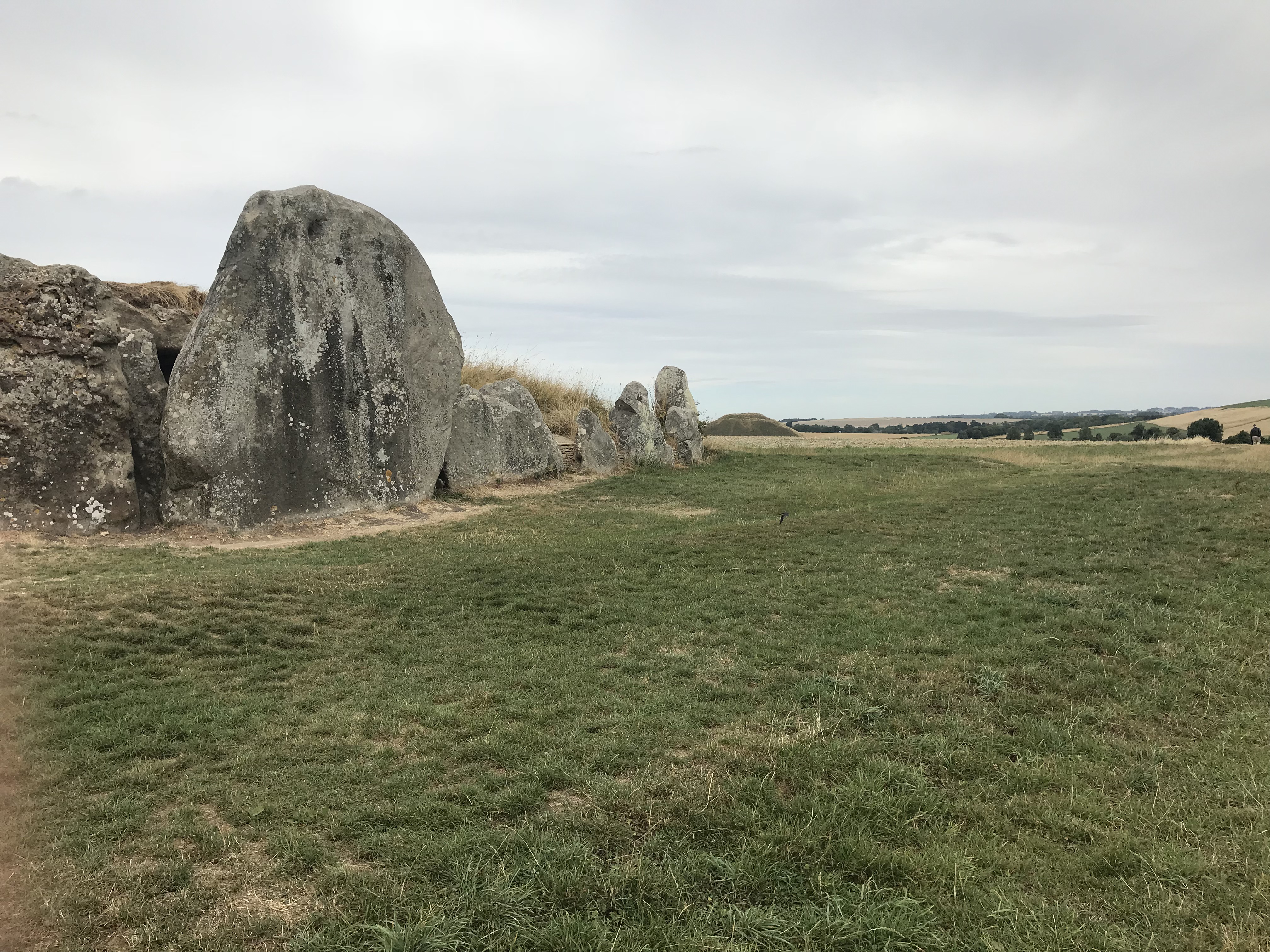 Upright stones edging a barrow to the left.  Beyond a bump rises on the horizon.