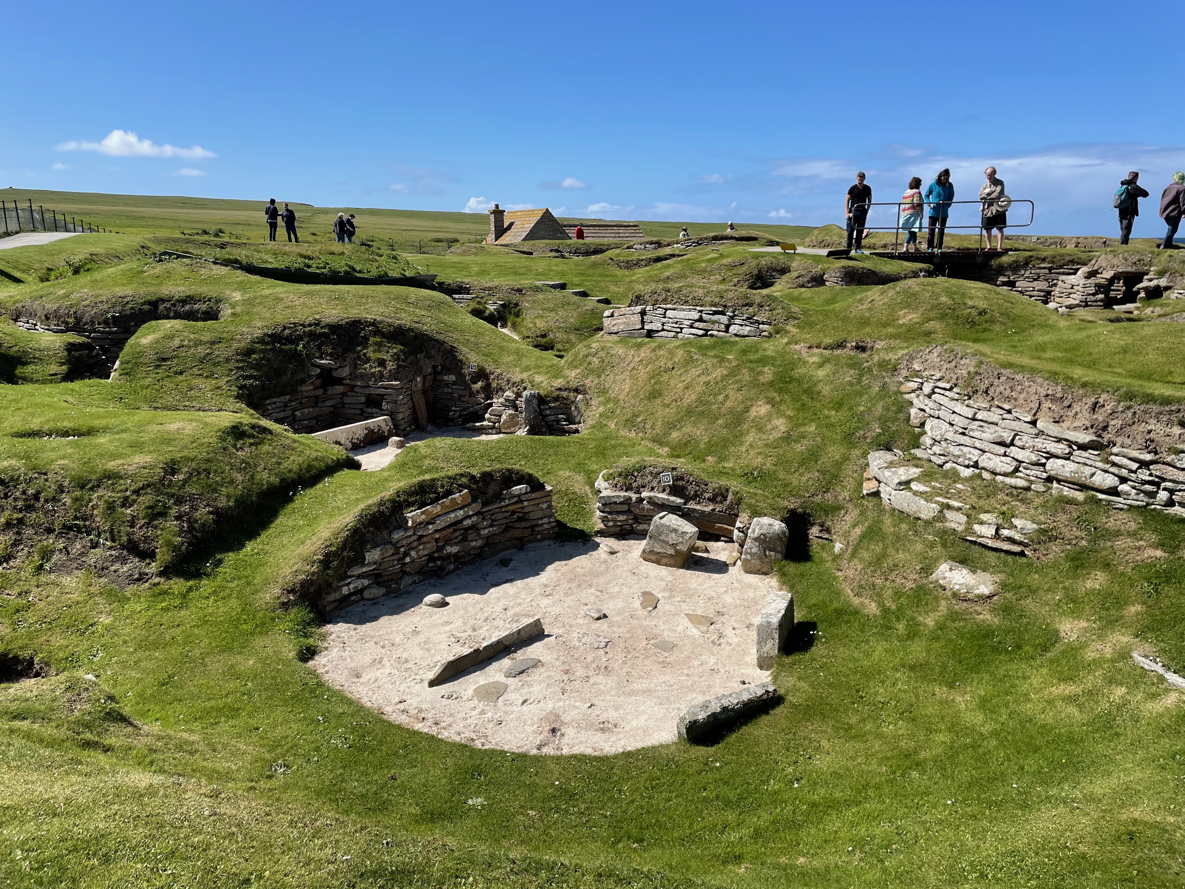 A circular hut exacted into the turf.  Some walls and a stones visible.  Other circular excavated dwellings visible.  Tourists against the skyline.