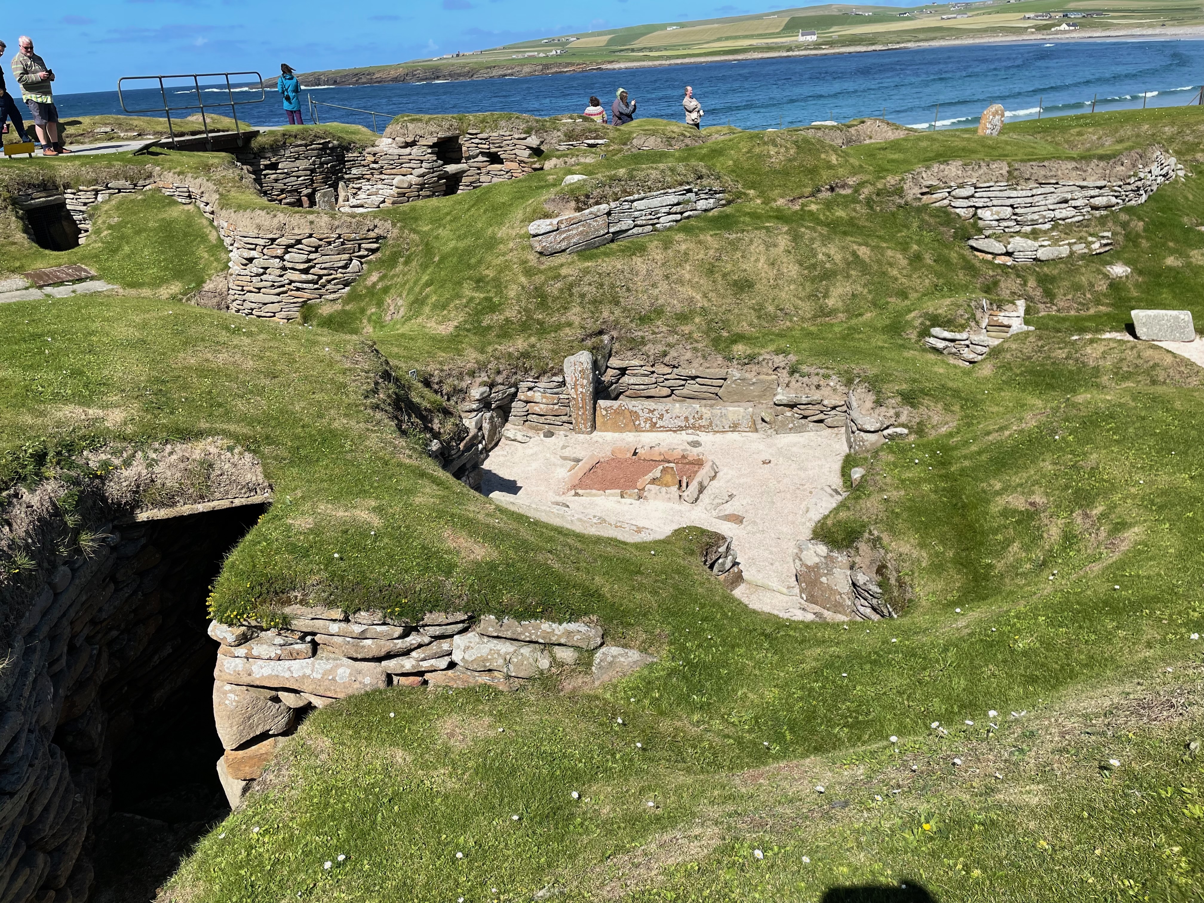View down from grassy mounds/walls into a neolithic hut.  There is a square hearth.  The walls of other huts are visible, sunken into the earth.  Tunnels can also be seen.  Beyond is the blue sea.
