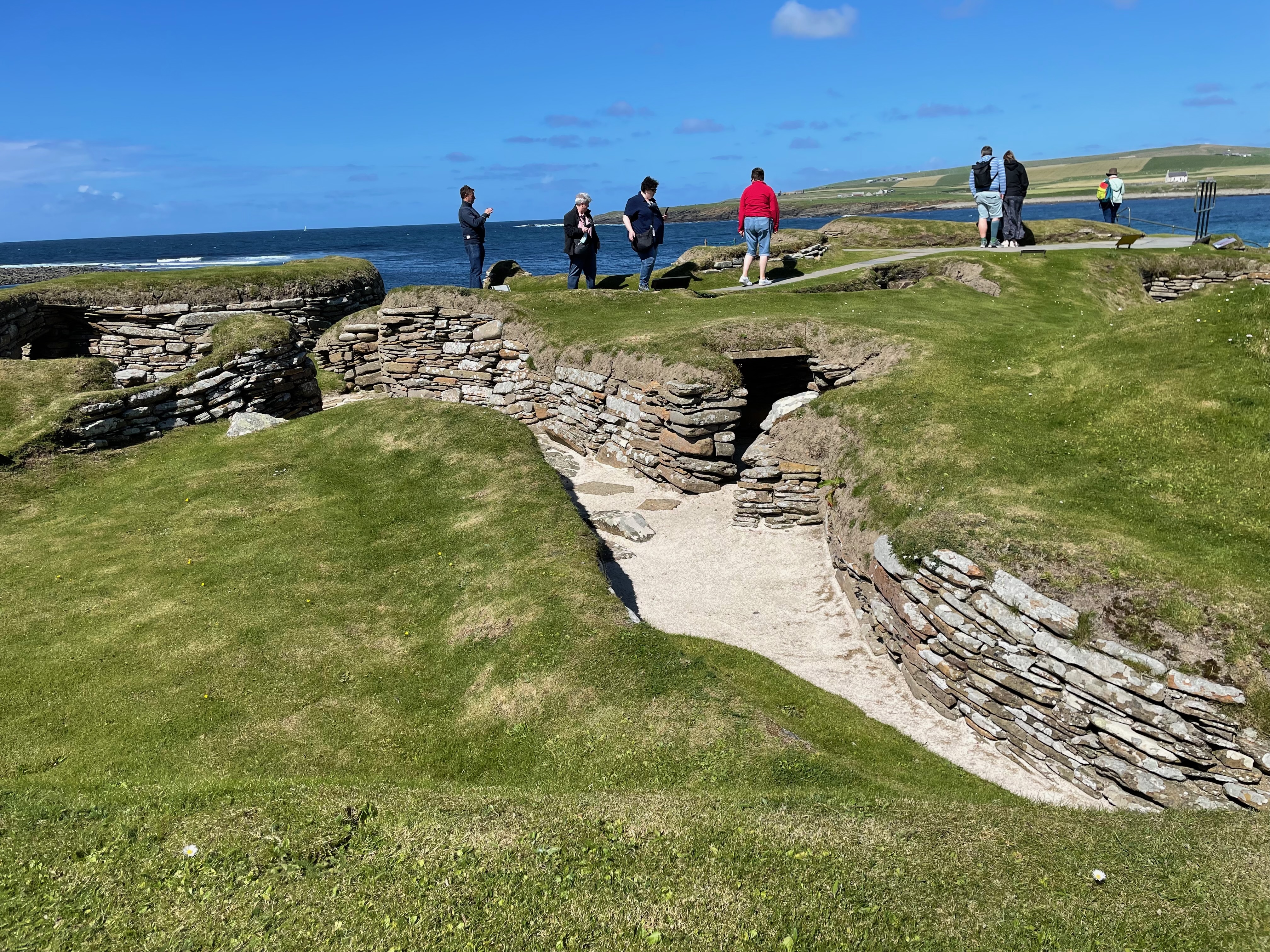 People standing on top of a grassy dunes, within which are walls and doorways.  There is blue sea and blue sky in the background.