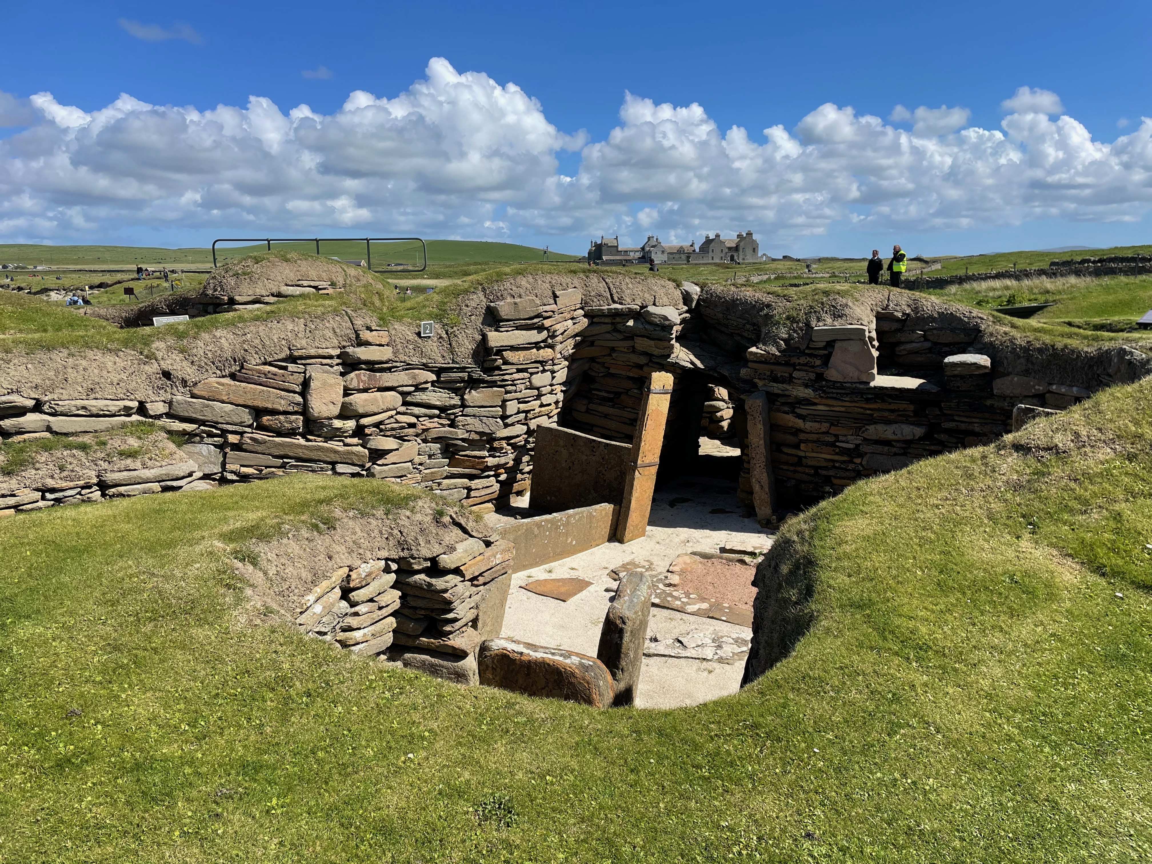Stone walls sunken into the ground, revealing rown rooms with a sandy floor.