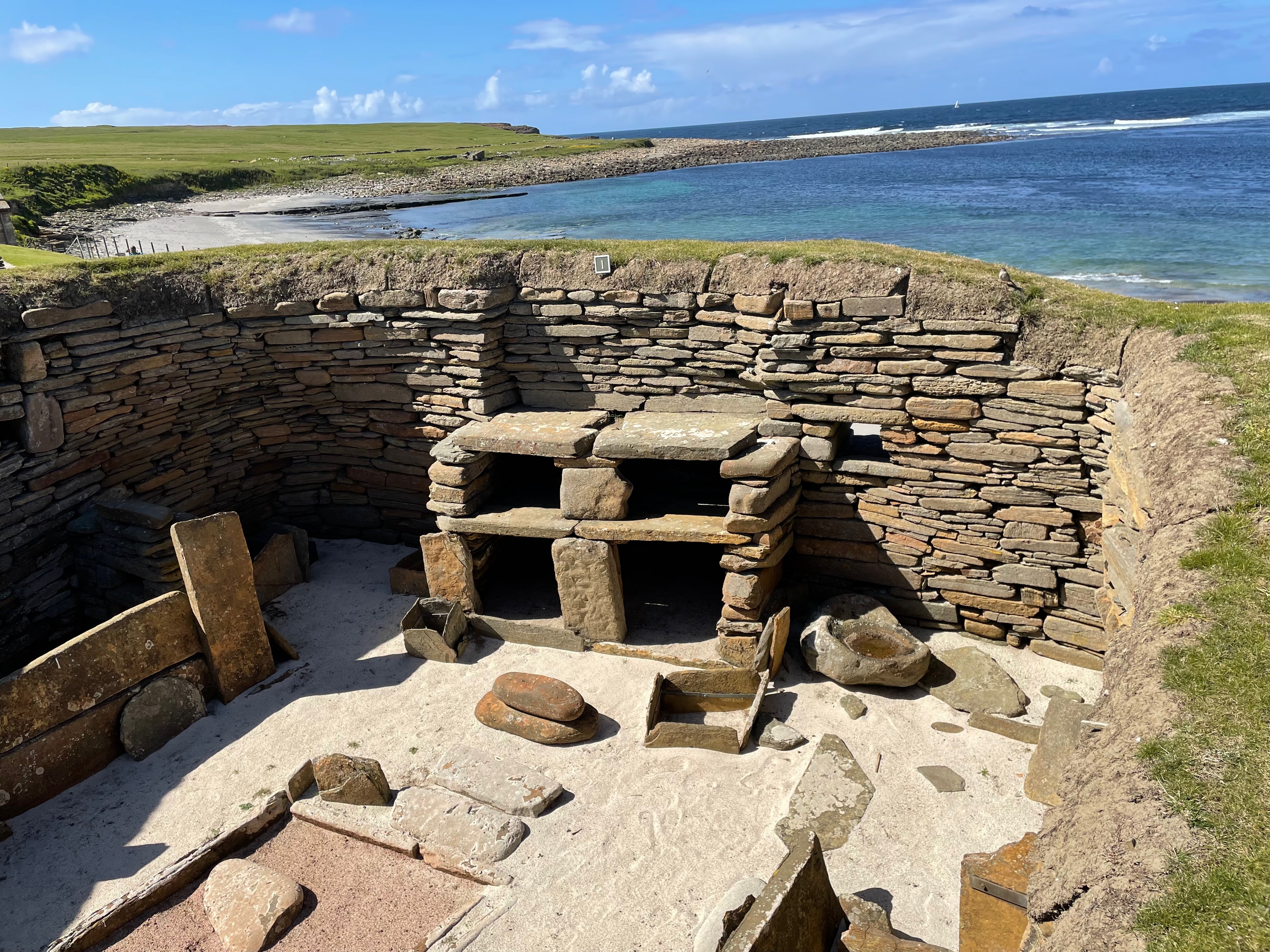 Looking down into a the remains of a stone hut in a sand dune. A stone dresser is prominent against one wall. Sea beyond.
