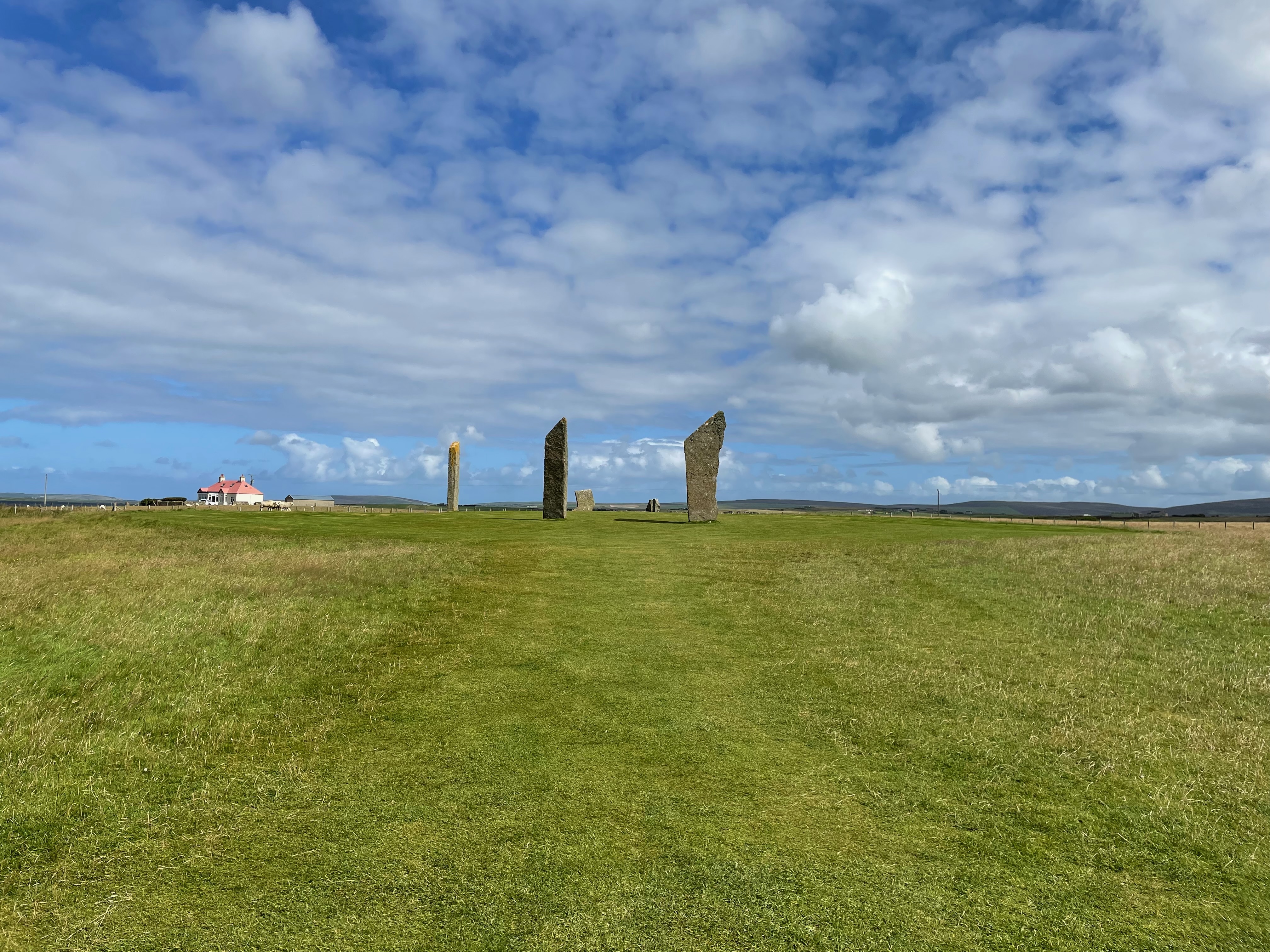 Wide flat grass and a blue sky with white clouds.  A white cottage is visible on the horizon to the right.  There are low hills in the far background.  Two tall sharp pointed stones stand at the end of a mown grass walk.  A further thin stone is visible sidways on between them and the cottage.  The low remains of a further stone are beyond.