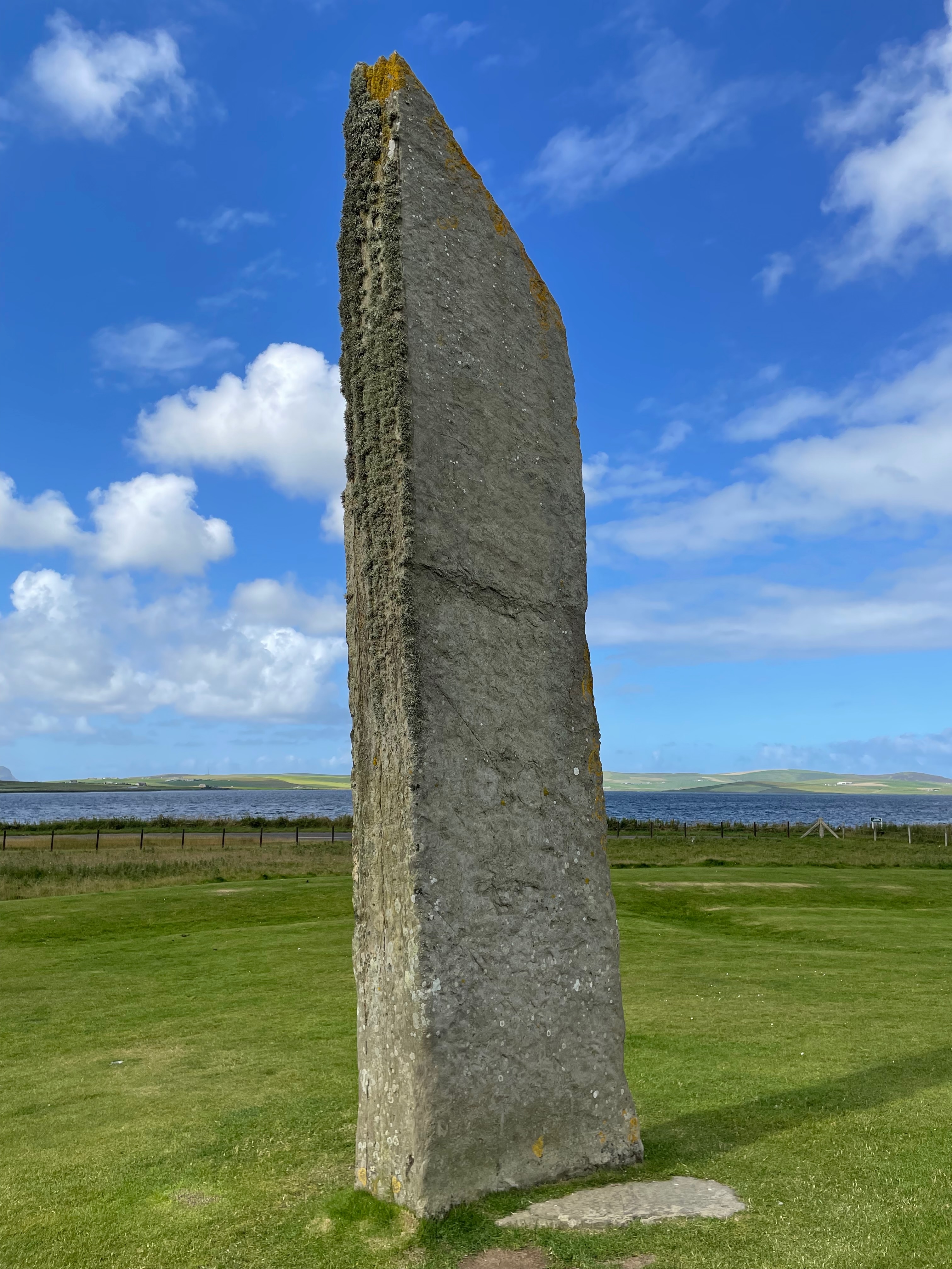 A tall thin standing stone with flat sides and a point.  Against a blue sky and a lake.