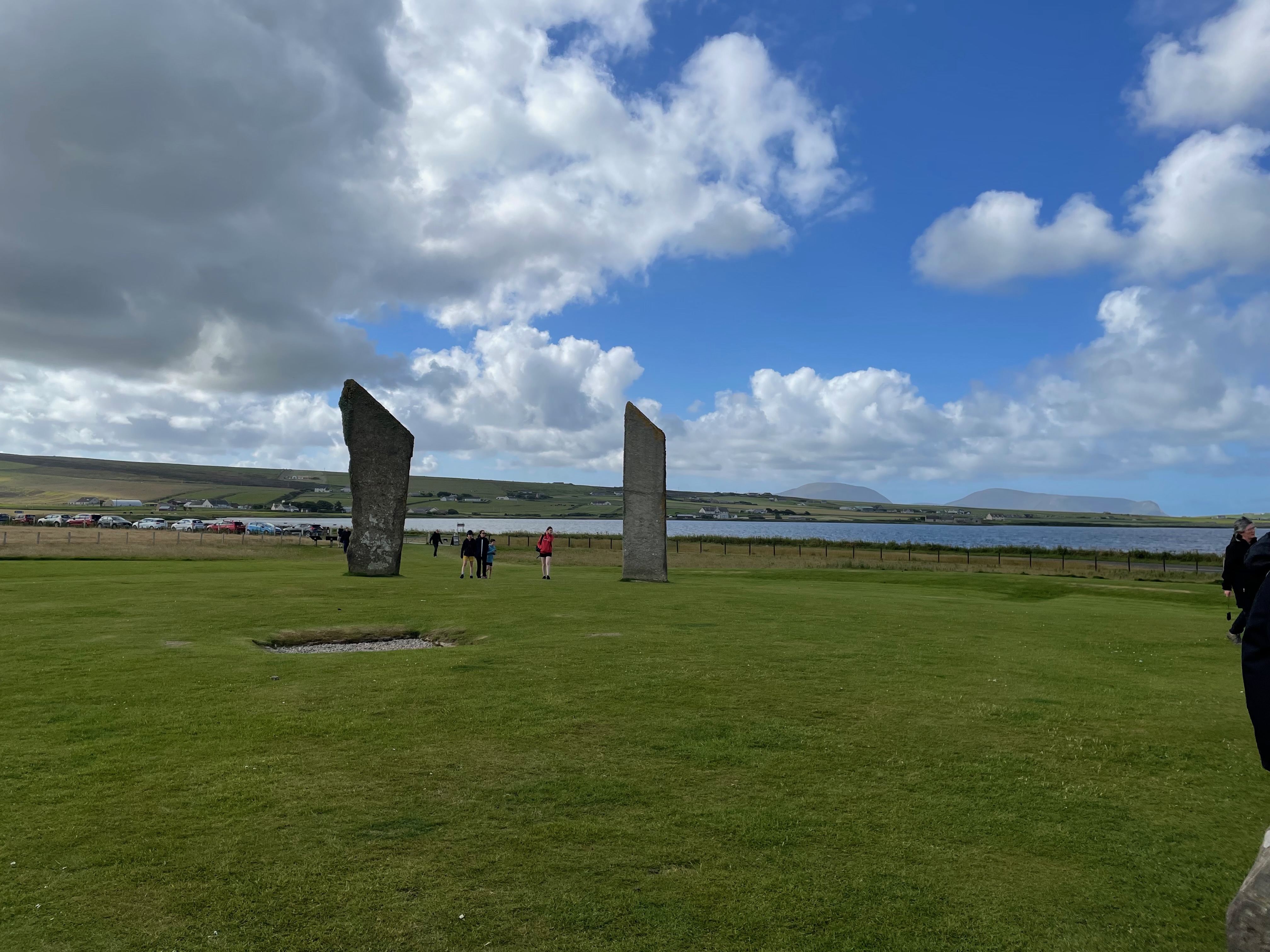 Two upright standing stones with people between them.  Low hills and a lake are in the background.