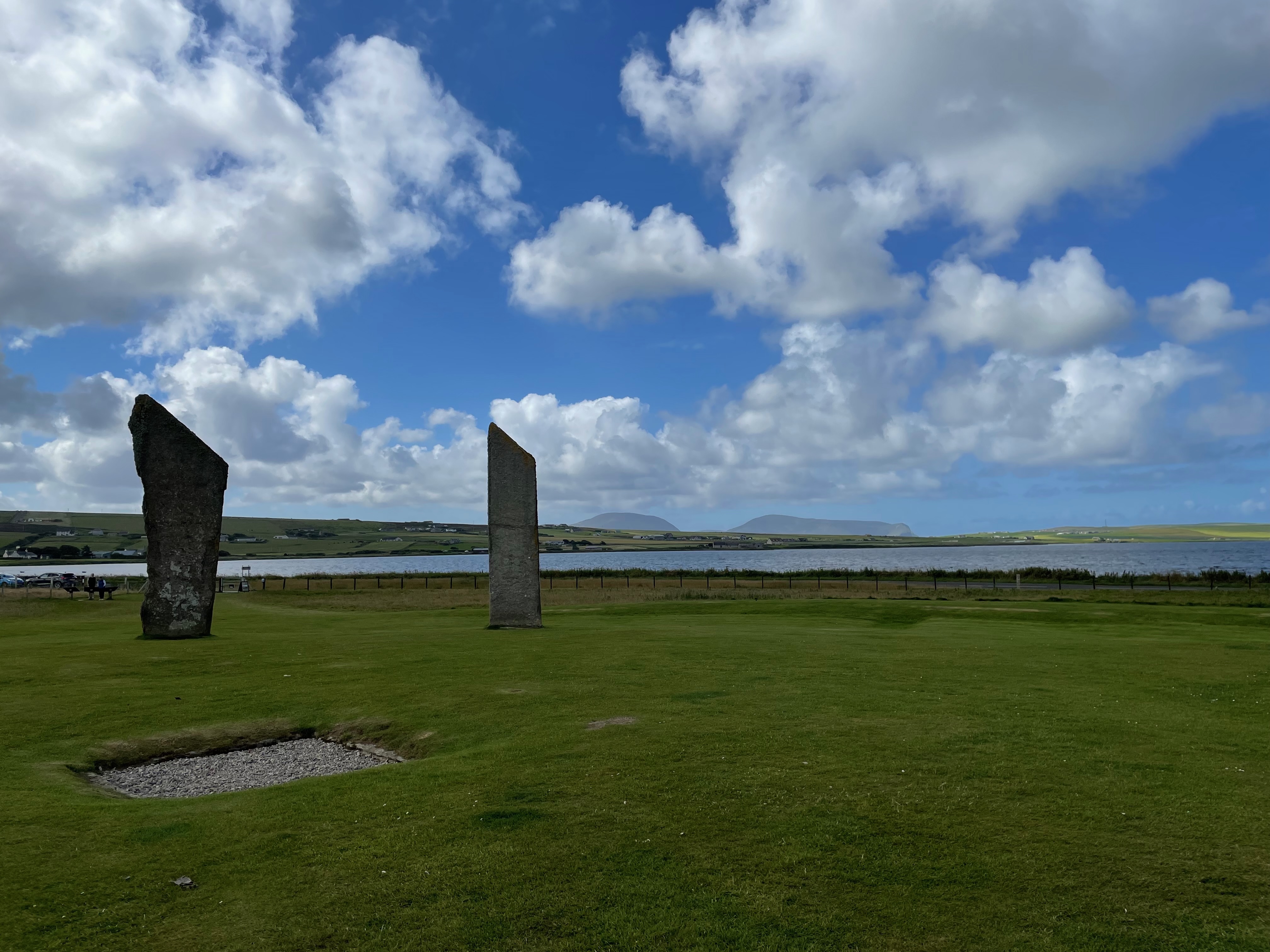 Two tall thin sharp standing stones in a grass area with a lake and sky beyond.