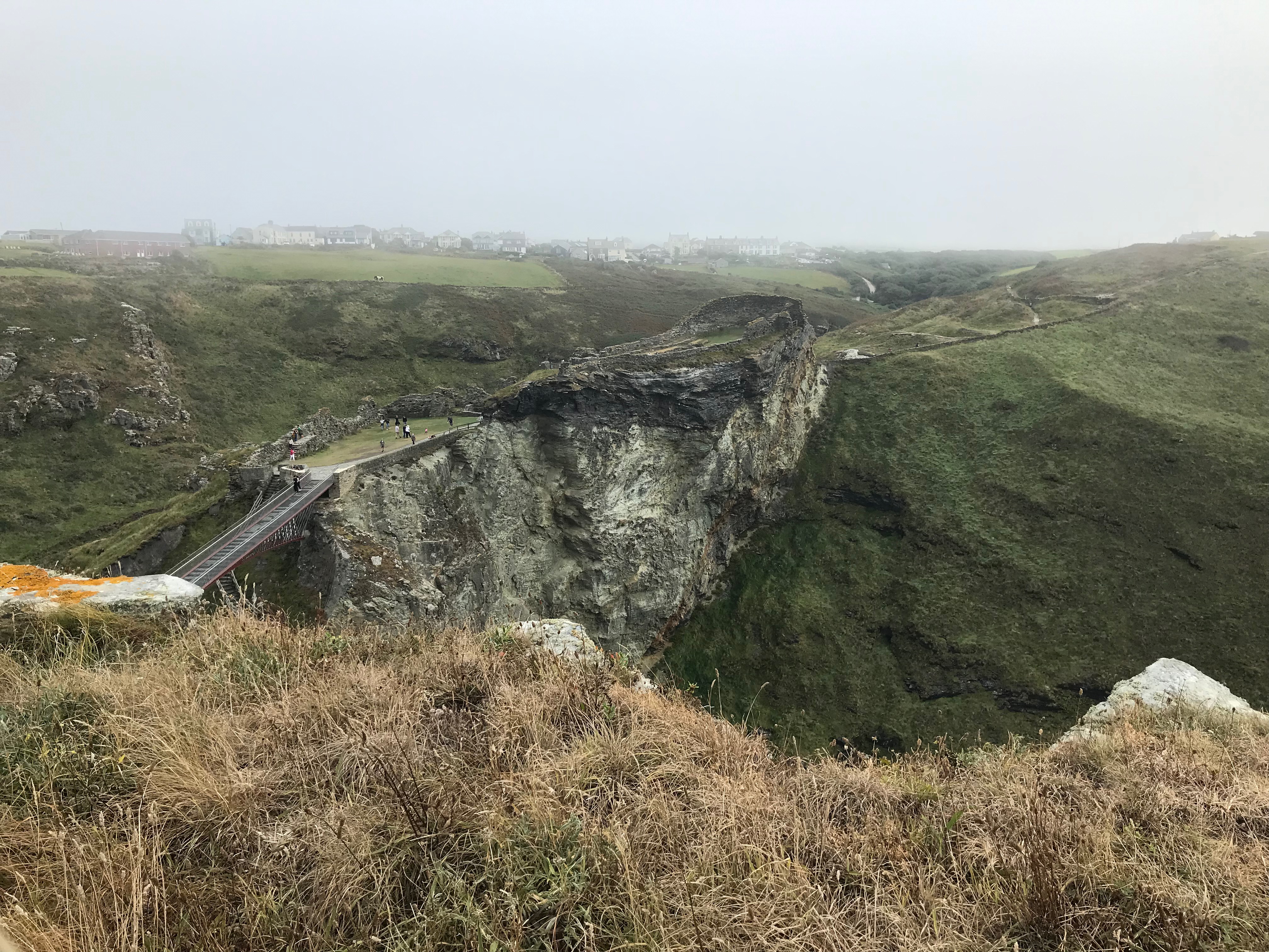 View across a bridged gap to the ruins of part of Tintagel castle on a cliff top.