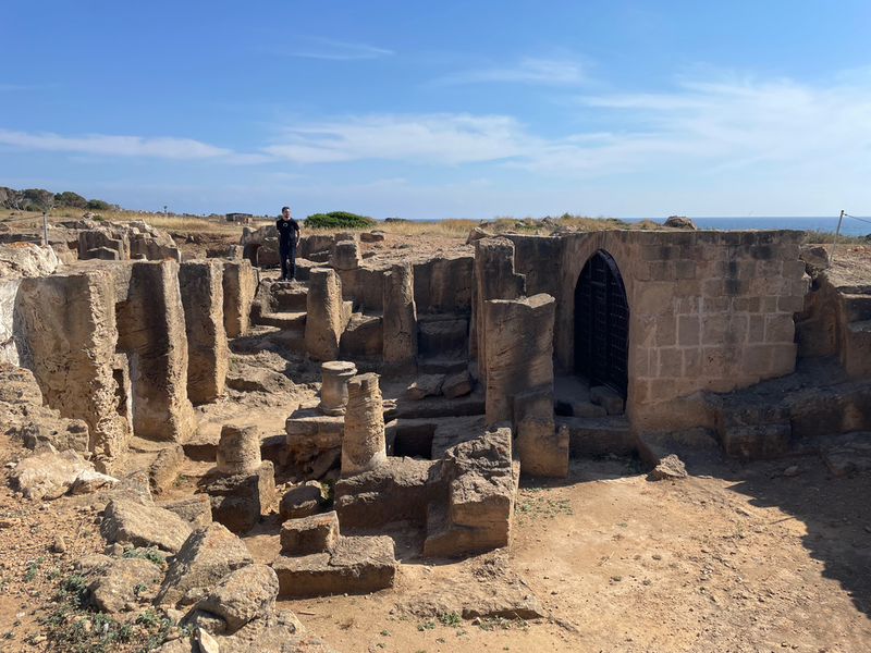 View of a building recessed into the ground. There are the low remains of circular pillars. Taller square pillars reaching up to ground level, and the arched entrance to a room. A blue sky above and a small amount of sea to the right. I man stands on wide steps leading up to ground level.