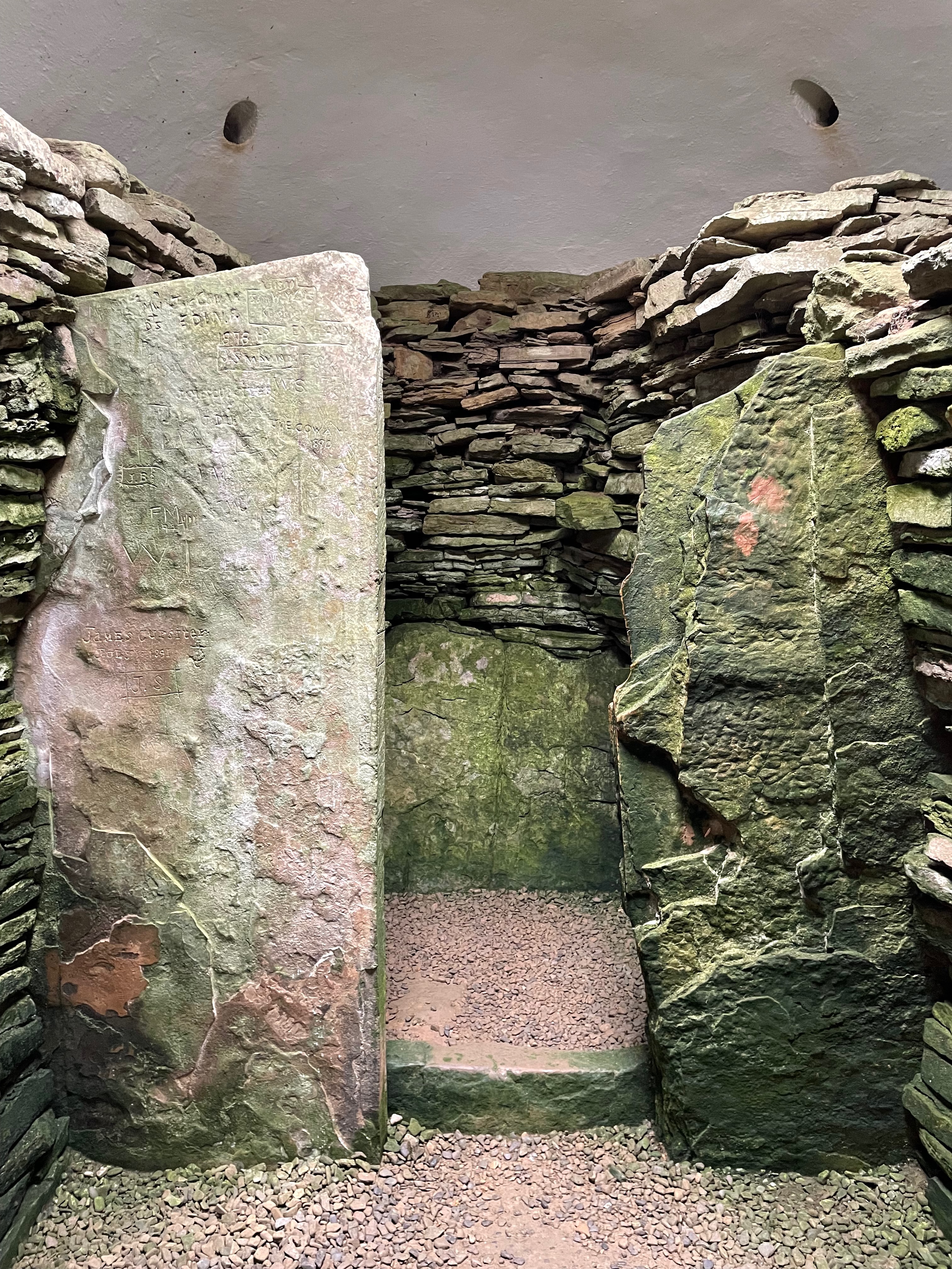 A small stone built chamber with a modern plastered roof.  Two upright stones stand sentinel against the walls creating a smaller chamber inside the first.  A stone step up and another low stone against the wall at the back create the impression of a bed or chair.