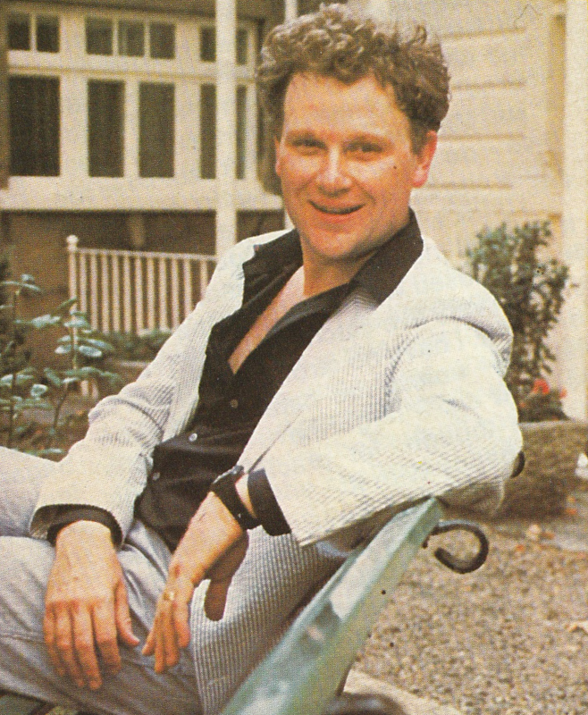 Colin Baker sitting on a garden bench. This is when he was cast as the doctor and his hair is shorter than it was in the show. He is wearing pale jeans and a black shirt with a blue and white striped jacket over it.