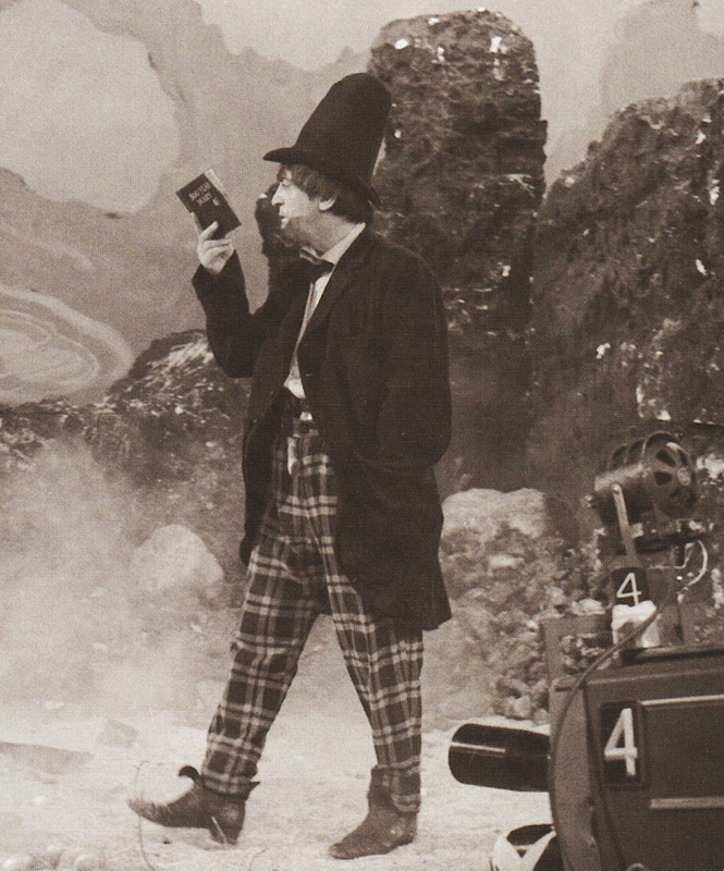 Black and White photo of Patrick Troughton as the Second Doctor. He is wearing a tall stove pipe hat and reading a small book as he strolls through an alien landscape, one hand in his pocket. There is a rocky backdrop and faint mist on the ground. A camera, numbered 4, is visible in the lower left.