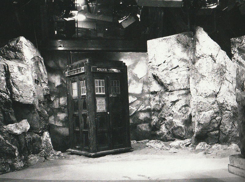 A black and white phot of a set containing flats representing rock faces and the Tardis.  Above the top of the flats studio lights can be seen.