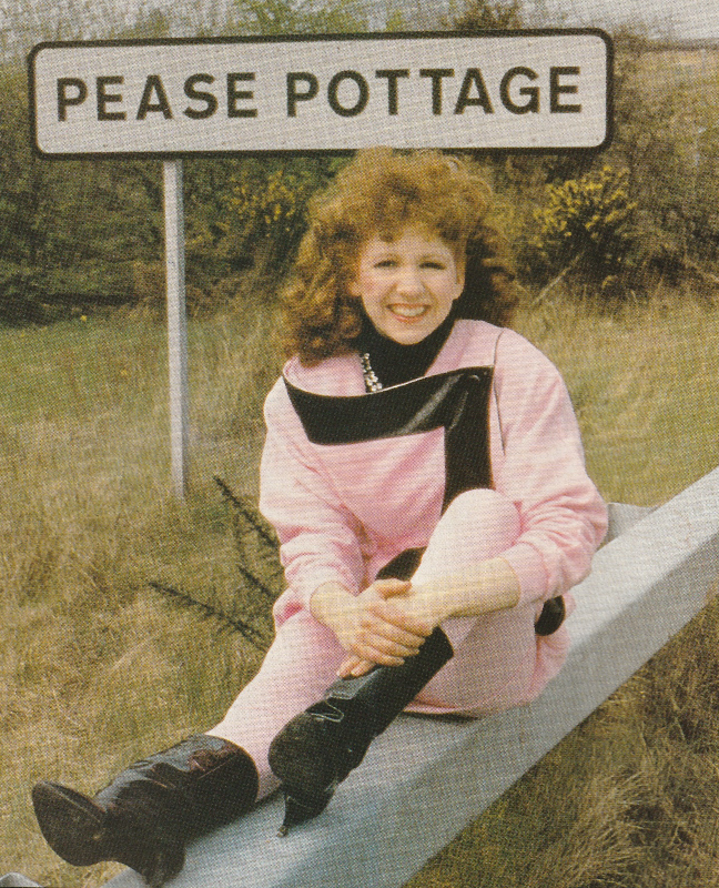 Bonnie Langford sitting on a motorway crash barrier in front of a Pease Pottage sign.  She is wearing a pink top and trousers with black high heeled boots.  The top fastens across the front with wide black edging.