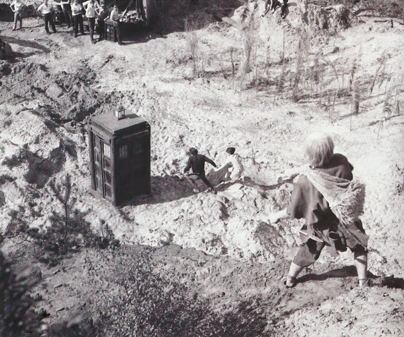 Black and white photo of Steven and Dodo fleeing to the Tardis, watched from above by a many in primitive clothing.  The film crew with a small tardis model can be seend in the top left corner.