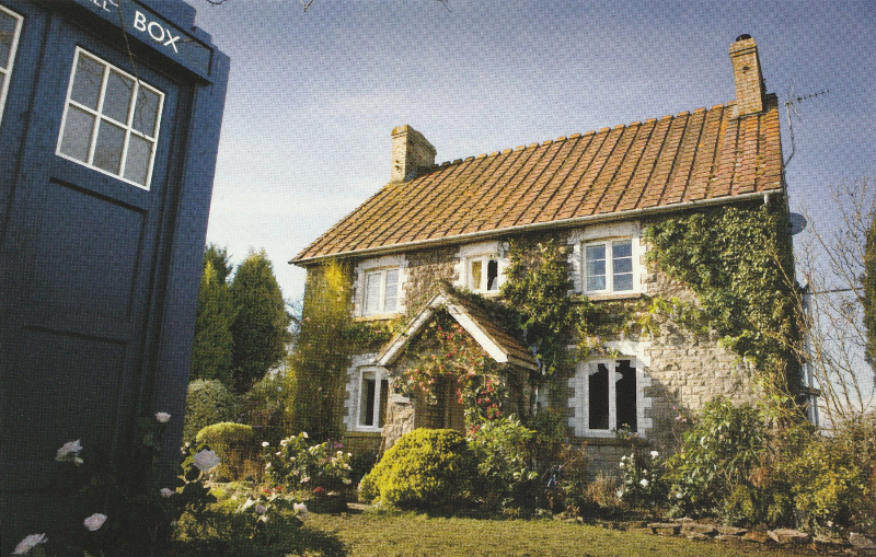 An ivy and rose covered house with a large front porch in the middle and five large windows, three on the top floor and two on the ground floor.  The tardis is at the edge of the image parked in the front garden.