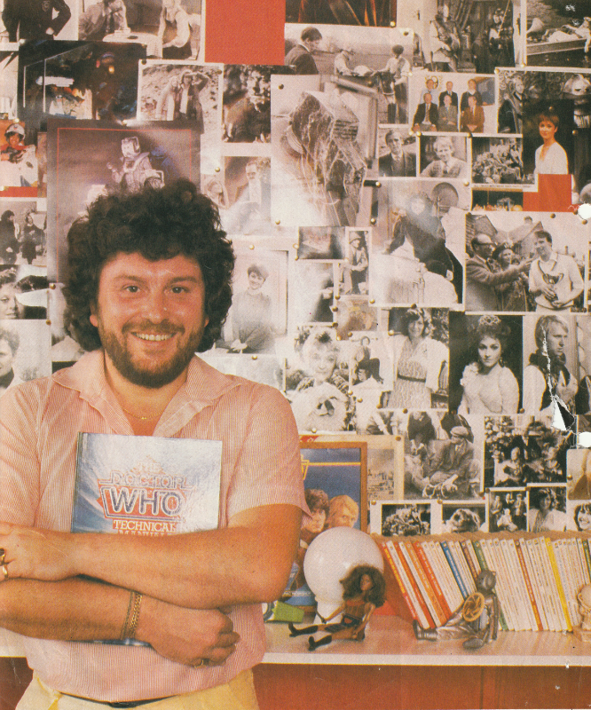 John Nathan-Turner holding a copy of the Doctor Who Technial Manual, standing in front of a wall of publicity photos from his time in Doctor Who, plus a few from All Creatures Great and Small.  On a shelf, below, are some Target Novelisations and a Leela and Cyberman doll.