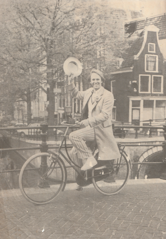Black and White publicity photo of Peter Davison dressed as the fifth Doctor riding a bicycle over a canal in Amsterdam.  He is waving his hat in one hand and looking at the camera.